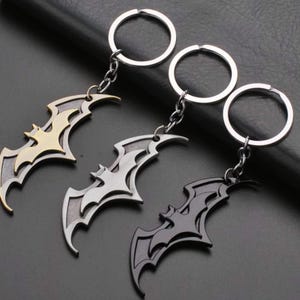 May include: Three metal bat-shaped keyrings in gold, silver, and black. Each keyring features a detailed bat silhouette design, connected to a chain and a silver ring. The keyrings are displayed on a dark surface.
