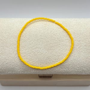 May include: A delicate yellow beaded necklace. The necklace is made of small, round beads strung together. The bright yellow color of the beads creates a cheerful and vibrant look. The necklace is displayed on a neutral-toned surface.