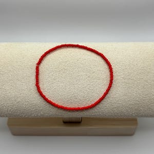 May include: A delicate red beaded bracelet, displayed on a neutral-toned surface. The bracelet is a continuous circle of small, vibrant red beads. The background is a light, solid color, highlighting the jewelry.