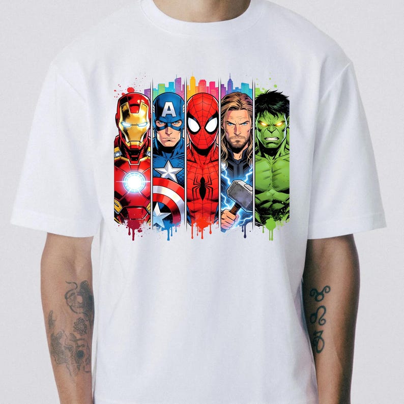 Op de afbeelding: Wit T-shirt met een kleurrijke afbeelding van vijf Marvel-superhelden: Iron Man, Captain America, Spider-Man, Thor en Hulk. Elk personage wordt afgebeeld in een verticaal paneel met een stadsgezicht op de achtergrond en een verfeffect.