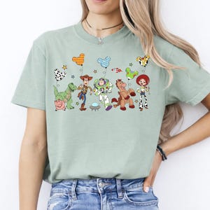 May include: A light green t-shirt featuring a colorful graphic of characters from the animated movie Toy Story. The design includes Woody, Buzz Lightyear, Jessie, and other characters, along with themed balloons. The shirt is made of a soft material.