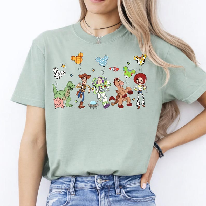 May include: A light green t-shirt featuring a colorful graphic of characters from the animated movie Toy Story. The design includes Woody, Buzz Lightyear, Jessie, and other characters, along with themed balloons. The shirt is made of a soft material.