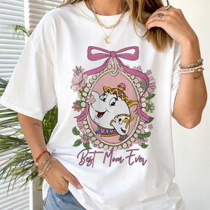 以下が含まれることがあります： 白のTシャツ。ピンクと金のフレームに美女と野獣のポット夫人とチップの画像、花とピンクのリボンで囲まれています。「Best Mom Ever」のテキストが画像の下に筆記体で書かれています。
