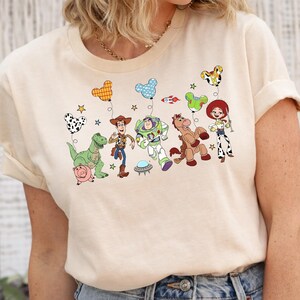 May include: A beige t-shirt featuring a colorful graphic of characters from the Toy Story movie. The design includes cartoon figures, balloons, and stars. The shirt has short sleeves and a crew neck.