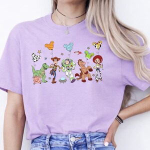 May include: A lilac-colored t-shirt featuring a colorful graphic of characters from the animated movie "Toy Story." The design includes Woody, Buzz Lightyear, Jessie, and other characters, along with balloons and stars. The shirt is a crew neck style.
