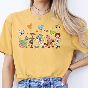 May include: Mustard yellow t-shirt featuring a colorful graphic of Toy Story characters. The design includes Woody, Buzz Lightyear, Jessie, and other characters with balloons. The shirt is a casual, comfortable style.