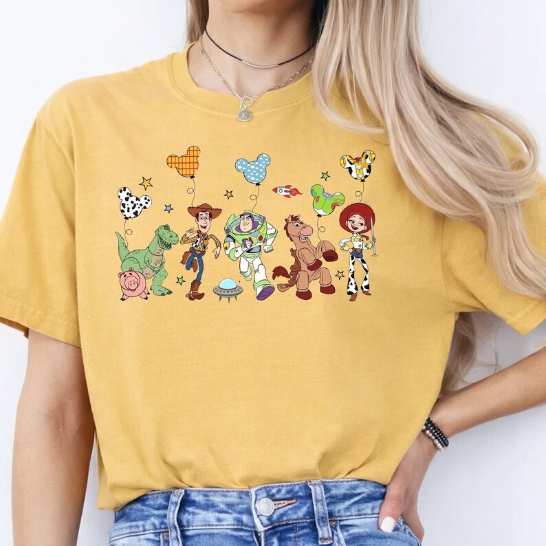 May include: Mustard yellow t-shirt featuring a colorful graphic of Toy Story characters. The design includes Woody, Buzz Lightyear, Jessie, and other characters with balloons. The shirt is a casual, comfortable style.
