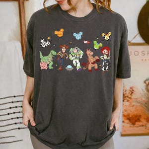 May include: Dark gray t-shirt featuring a colorful graphic of characters from the Toy Story movie. The design includes Woody, Buzz Lightyear, and other characters, along with decorative elements. The shirt is a casual wear item.