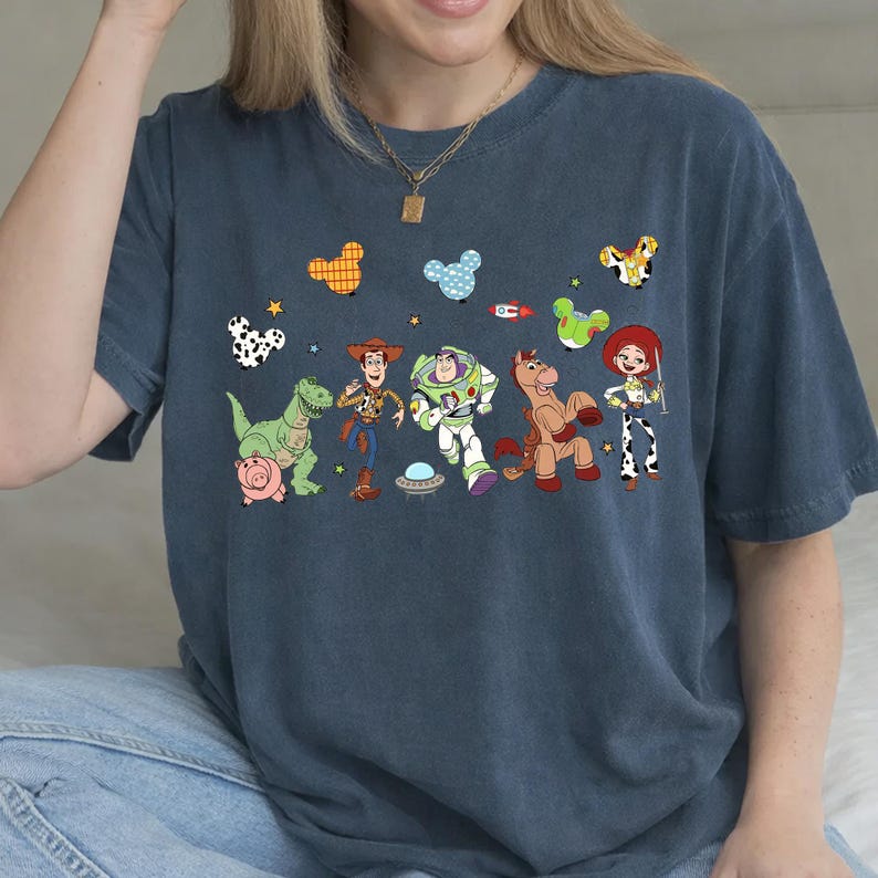 May include: A navy blue t-shirt featuring a colorful graphic of characters from the animated movie "Toy Story." The design includes Woody, Buzz Lightyear, Jessie, and other characters, along with decorative shapes. The shirt is made of a soft material.