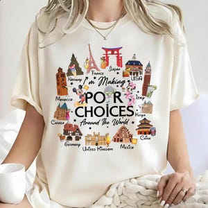 May include: Cream-colored t-shirt with a colorful graphic. The graphic features illustrations of landmarks from around the world, including the Eiffel Tower and the Taj Mahal, with the text "I'm Making Poor Choices Around The World."