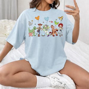 May include: Light blue t-shirt featuring a colorful graphic of Toy Story characters and balloons. The design includes Woody, Buzz Lightyear, and other characters from the animated film. A fun and playful design for fans of the movie.