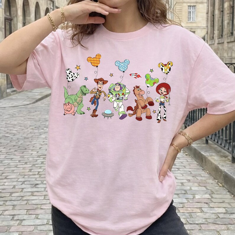 May include: A light pink t-shirt featuring a colorful graphic of characters from the animated movie Toy Story. The design includes Woody, Buzz Lightyear, and other characters, along with balloon-shaped designs. The shirt is a crew neck style.