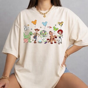May include: Cream-colored t-shirt featuring a colorful graphic of animated characters. The design includes a dinosaur, a pig, Woody, Buzz Lightyear, a horse, and Jessie. Balloons and stars are also part of the design.