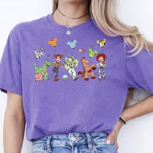 May include: A purple t-shirt featuring a colorful graphic of characters from the animated movie Toy Story. The design includes Woody, Buzz Lightyear, Jessie, and other characters. The shirt is made of a soft material.