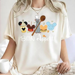 Puede incluir: Camiseta color crema con una ilustración de dibujos animados de una rata chef sosteniendo una cuchara de madera, rodeada de alimentos como helado, manzanas de caramelo, una hamburguesa y algodón de azúcar. El texto "Bon Appétit!" está escrito en blanco.