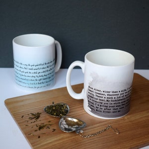 May include: Two white ceramic mugs with black text and watercolor designs. The mug on the right has the text "Moon River, wider than a mile, I'm crossing you in style someday..." The mugs are on a wooden surface with a heart-shaped tea infuser.