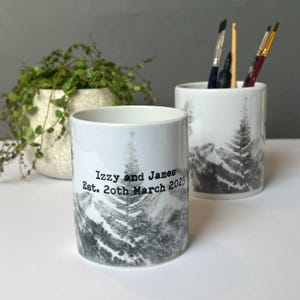 May include: Two white ceramic pencil holders with a black and gray forest design. One holder has the text "Izzy and James Est. 20th March 2025". The other holder contains paint brushes and a crochet hook. A potted plant is in the background.