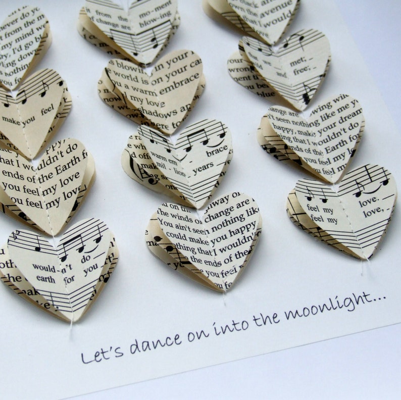 May include: A white card with the text "Let's dance on into the moonlight..." written in black ink. The card is decorated with a string of paper hearts made from music sheets. The hearts are arranged in a line, with the text at the bottom of the card.