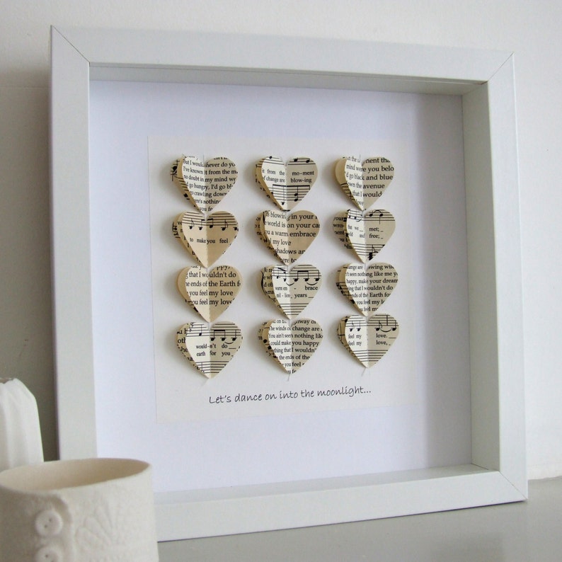 May include: A white frame with a white mat surrounding a collection of paper hearts made from sheet music. The hearts are arranged in a vertical line and are all facing the same direction. The text "Let's dance on into the moonlight..." is printed below the hearts.