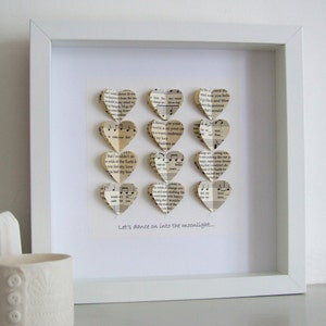 May include: A white frame with a white mat and a white background. The frame contains a collection of paper hearts made from sheet music. The hearts are arranged in a vertical row. The text "Let's dance on into the moonlight..." is printed below the hearts.