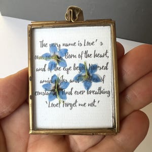 May include: A rectangular brass-framed display with a white background, featuring blue forget-me-not flowers and handwritten text. The text reads, "The very name is Love's...Love! Forget me not."