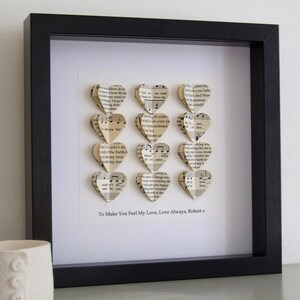 May include: A framed piece of art with a white background and a black frame. The art features a series of paper hearts made from sheet music, arranged in a vertical column. The text "To Make You Feel My Love, Love Always, Robert x" is printed below the hearts.