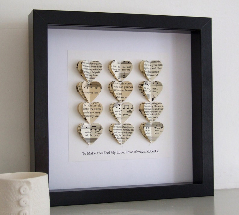 May include: A framed piece of art with a white background and a black frame. The art features a series of paper hearts made from sheet music, arranged in a vertical column. The text "To Make You Feel My Love, Love Always, Robert x" is printed below the hearts.