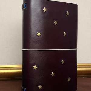 Könnte beinhalten: Ein dunkelburgunderfarbenes Lederjournal mit goldenen Sternverzierungen. Das Journal wird mit einem weißen Gummiband verschlossen und hat blaue Nähte am Buchrücken. Der Einband ist glatt und die Sterne sind gleichmäßig verteilt.