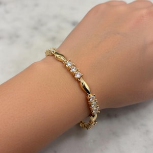 Könnte beinhalten: Ein goldfarbenes Armband mit klaren, funkelnden Steinen. Das Armband hat ovale Glieder, die sich mit Abschnitten aus mehreren kleinen Steinen abwechseln. Das Armband wird am Handgelenk getragen und zeigt sein Design und seinen Glanz.
