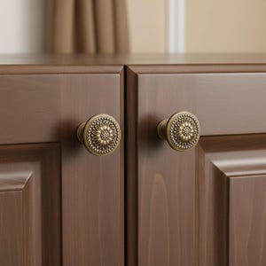 Peut inclure: Deux boutons de placard ronds avec une finition en laiton antique, présentant un motif de pétales de tournesol ou de marguerite en relief au centre, entouré d'une bordure perlée, fixés sur des portes d'armoire en bois brun foncé.