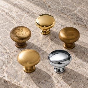 May include: Five round cabinet knobs arranged in a circle. One knob has a polished gold finish, one is brushed gold, one is a textured antique gold, one is a brushed bronze, and one is a polished chrome finish. Each knob has a central stem base.