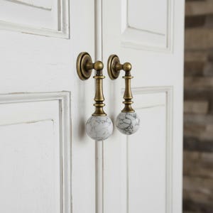 May include: Two antique brass cabinet pulls with white and gray marble sphere knobs. The pulls are mounted on a white paneled door. The hardware has a vintage aesthetic.
