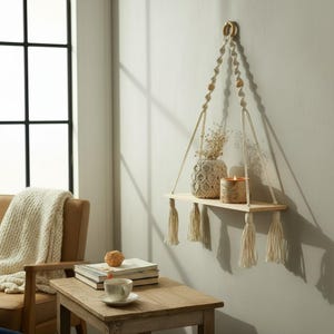 Puede incluir: Una estantería colgante de madera con diseño macramé. La estantería está suspendida por una cuerda y adornada con borlas. Contiene un jarrón decorativo, una vela y flores secas. La estantería está montada en una pared gris claro.