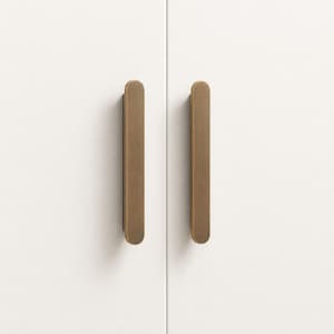 May include: Two vertical, brushed brass cabinet pulls with a rounded, elongated rectangular shape. These metallic handles are mounted side-by-side on a smooth, matte white surface, featuring a consistent finish and a clean, simple design.