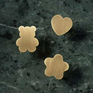 Può includere: Tre pomelli per mobili in metallo con finitura oro spazzolato. Il set include un pomello a forma di orsetto, uno a forma di cuore e uno a forma di fiore a cinque petali. Ogni pezzo è caratterizzato da una superficie liscia e piatta.