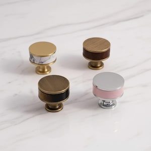May include: Four cylindrical cabinet knobs with distinct finishes. One features a gold top with a white marble band, one has a wood grain top with a dark brown band, one is brushed brass with a black band, and one is silver with a light pink band.