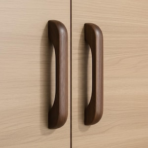 Può includere: Due maniglie per armadietto in legno marrone scuro con una forma liscia, curva e ad arco. Le maniglie sono montate verticalmente su una superficie di anta color beige chiaro con venature del legno, con una distinta giuntura verticale tra le ante.