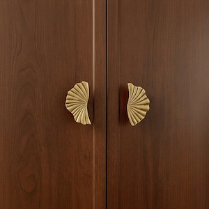May include: Close-up of a wooden cabinet with two gold-colored, fan-shaped handles. The cabinet doors are a dark brown color with a wood grain texture. The handles are symmetrical and add a decorative touch to the cabinet.