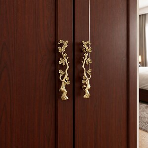 May include: Two vertical door handles made of polished gold-toned metal, each featuring a detailed sculptural design of a flowering tree branch with multiple small blossoms and a textured trunk, mounted onto a dark brown wood-grain cabinet door.