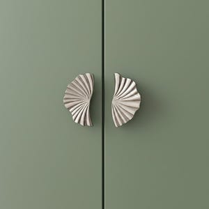May include: Two silver-toned metal cabinet pulls with a fan-shaped, ribbed design mounted on a matte sage green surface. The handles feature a curved, semi-circular silhouette with vertical grooves radiating from the center attachment point.