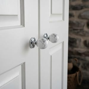 May include: White marble-style door knobs with chrome bases are mounted on a white door. The knobs have a spherical shape with gray veining, adding a touch of elegance to the door's design. The door has a paneled construction.