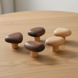 May include: Five wooden mushroom-shaped cabinet knobs. Two are a dark walnut color, and three are a light natural wood color. The knobs have a rounded top and a short, cylindrical base. They are arranged on a light brown wooden surface.
