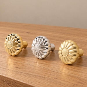 May include: Three round cabinet knobs with a flower-like petal design. The left knob is polished gold, the center knob is polished silver, and the right knob is a matte gold finish. Each features a textured center and a beaded border.