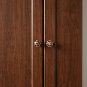 Puede incluir: Dos puertas de armario de madera de color marrón oscuro con un patrón de veta de madera visible. Cada puerta está equipada con un pomo redondo de metal color latón antiguo que muestra un patrón floral o de roseta detallado con un borde.