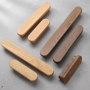 Può includere: Una collezione di maniglie per mobili in legno di varie dimensioni e tonalità di marrone. Le maniglie hanno una forma rettangolare arrotondata e sono realizzate in diversi tipi di legno, tra cui tonalità chiare e scure. L'hardware è disposto su una superficie grigia.