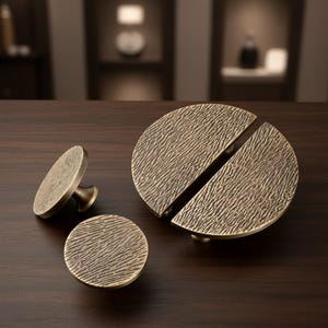 May include: A set of antique brass cabinet hardware, including a round knob, a small pull, and a large, divided circular pull. The hardware has a textured surface and is displayed on a dark wood surface.