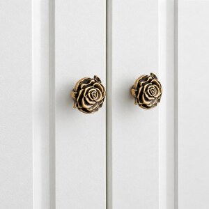 Peut inclure: Deux boutons de placard en métal couleur laiton antique en forme de roses épanouies. Chaque bouton présente des pétales détaillés et superposés sur une base circulaire, fixés sur une porte de placard blanche à panneaux verticaux.