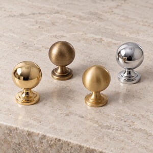 May include: Four spherical cabinet knobs with circular bases. One knob has a polished gold finish, one has a brushed antique brass finish, one has a matte satin brass finish, and one has a reflective polished chrome finish.