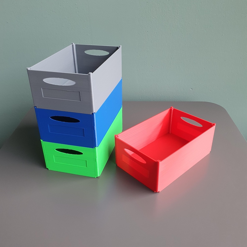 May include: A stack of three rectangular storage bins in gray, blue, and green, with a separate red bin. Each bin has a handle cutout. The bins are on a gray surface.