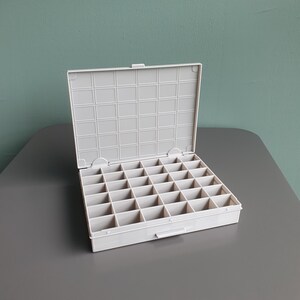 May include: A light grey plastic storage organiser box featuring a hinged lid and a grid of thirty-two individual square compartments. The interior base is divided into four rows of eight sections, and the lid interior has a matching grid pattern.